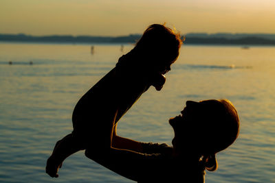Silhouette woman holding baby by sea against sky during sunset