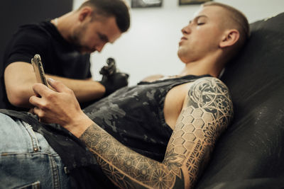 Tattooed man using smartphone during tattoo session, male artist draws on the clients skin	
