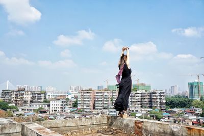 Side view of young woman with arms raised standing on retaining wall of building terrace