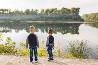 Rear view of boys standing by lake against trees
