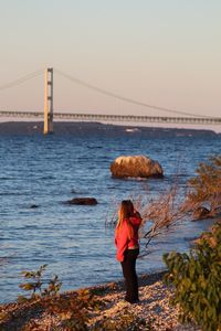 Side view of woman standing at beach with bridge in background during sunset