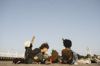 Teenage girls pointing at heart shape vapor trail on clear sky during sunny day