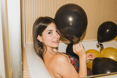 From above of young female in festive dress laying down in bathtub looking at camera filled with colorful balloons during holiday celebration