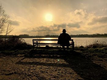 Rear view of man sitting by lake on bench during sunset