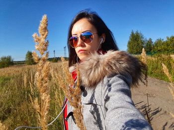 Portrait of young woman wearing sunglasses standing on field against clear blue sky