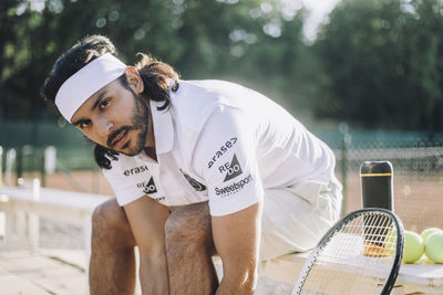 Confident man wearing headband while sitting on bench at tennis court