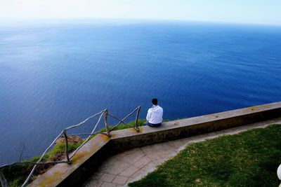 Rear view of man sitting on retaining wall against blue sea