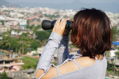 Side view of woman looking through binoculars against cityscape