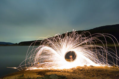 Man spinning illuminated wire wool by lake against sky at night