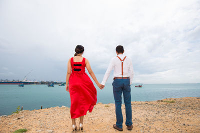 Rear view of couple holding hands while standing at beach against cloudy sky