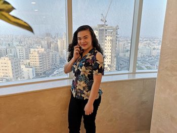 Full length of smiling young woman standing against window in city