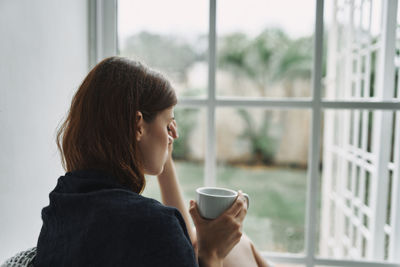 Depressed woman holding coffee cup sitting by window