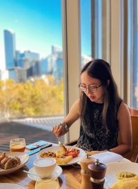 Young woman having breakfast at table against window view of high-rises in the city.