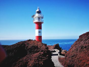 Lighthouse by sea against clear blue sky