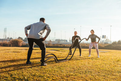 Women exercising with rope by instructor on land against sky in sunny day