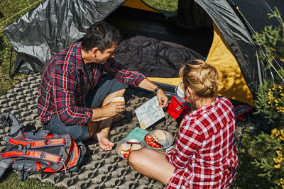 Couple having breakfast and planning their day sitting by tent at camping. people spending vacations