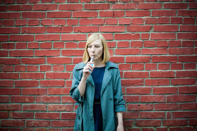 Beautiful young woman smoking electronic cigarette against brick wall