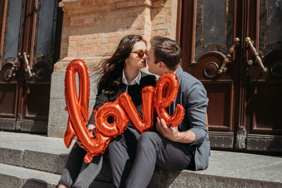 Couple holding balloon kissing while sitting outdoors