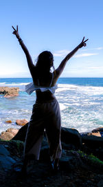 Rear view of young woman with arms raised standing at beach against sky