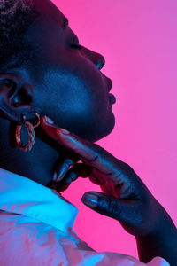 Side view of delicate african american female model in trendy earrings touching neck in studio with neon light on pink background