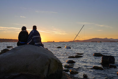 Couple on rock by sea against sky during sunset