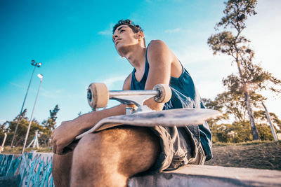 Low angle view of man with skateboard sitting on retaining wall against sky