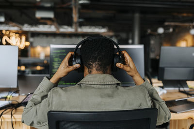 Rear view of male coder wearing wireless headphones sitting at desk in office