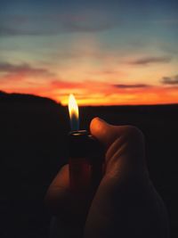 Cropped hand holding cigarette lighter against sky during sunset
