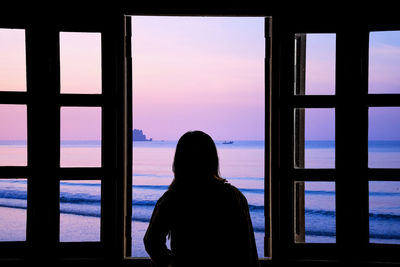 Rear view of silhouette woman looking through window at sunset