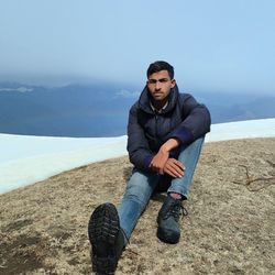 Portrait of young man sitting on rock against sky