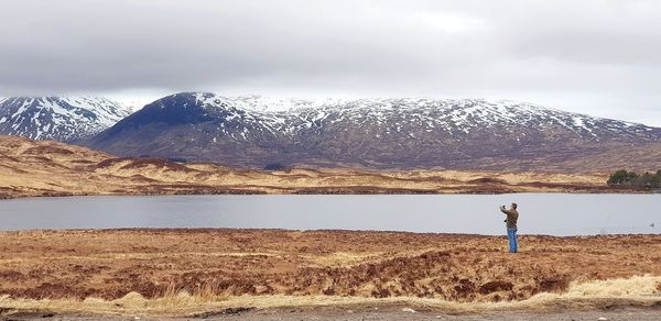 Distant view of man standing at lakeshore against mountains during winter