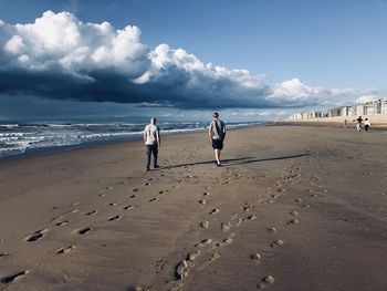 Rear view of 2 people walking at beach against sky