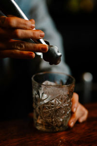 Crop female barkeeper putting ice cube with tongs into glass while preparing cocktail in bar with bottles on blurred background