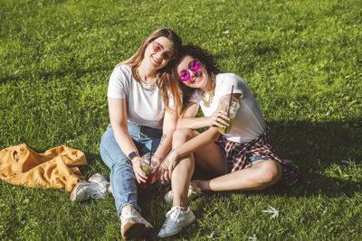 Happy female friends sitting with lemonade on grass at park during sunny day