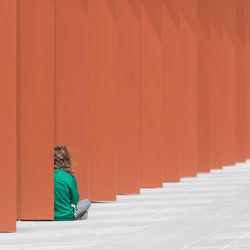 Side view of woman sitting against orange wall