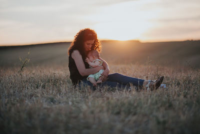 Woman with daughter sitting on grass against sky during sunset