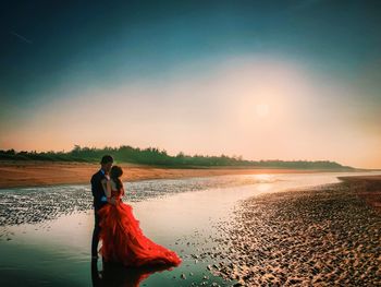 Full length of couple standing at beach against sky during sunset