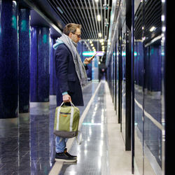 Full length of man using smart phone while standing in illuminated corridor