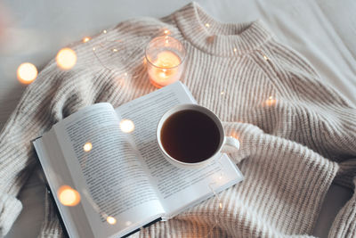 Open book with cup of coffee in bed with knit cloth sweater over glow christmas lights at night.