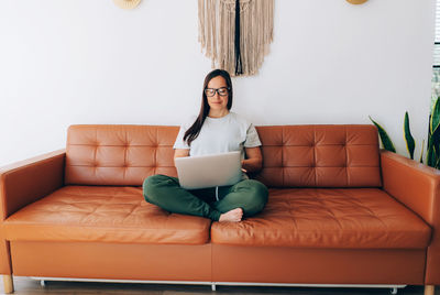Young smiling woman in glasses working on her laptop sitting on the couch at home
