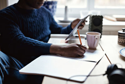 Midsection of teenage boy holding digital tablet while writing on book at home