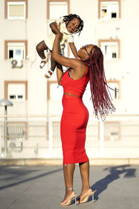 Side view of african american female with long braids in stylish outfit standing and lifting happy little daughter while having fun on street in sunny day