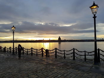 Silhouette people on street by sea against sky during sunset