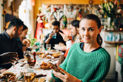 Portrait of confident young woman holding smart phone while sitting with friends at table in restaurant during dinner pa