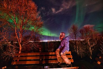 Side view of person sitting on bench against northern lights at night