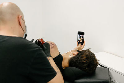 Content female in protective respirator lying on table and taking photo on smartphone while getting tattoo in salon with male master