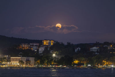 Illuminated buildings by sea against sky at night