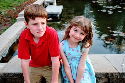 Siblings sitting on retaining wall against pond