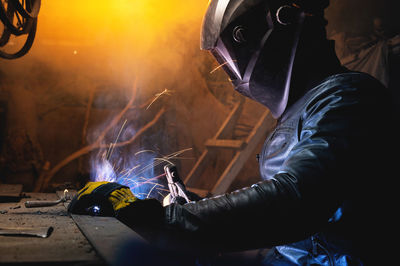 A male welder in a leather jacket carries out welding work in a home shed against the background of