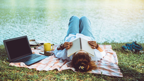 Woman with book lying by laptop at lakeshore